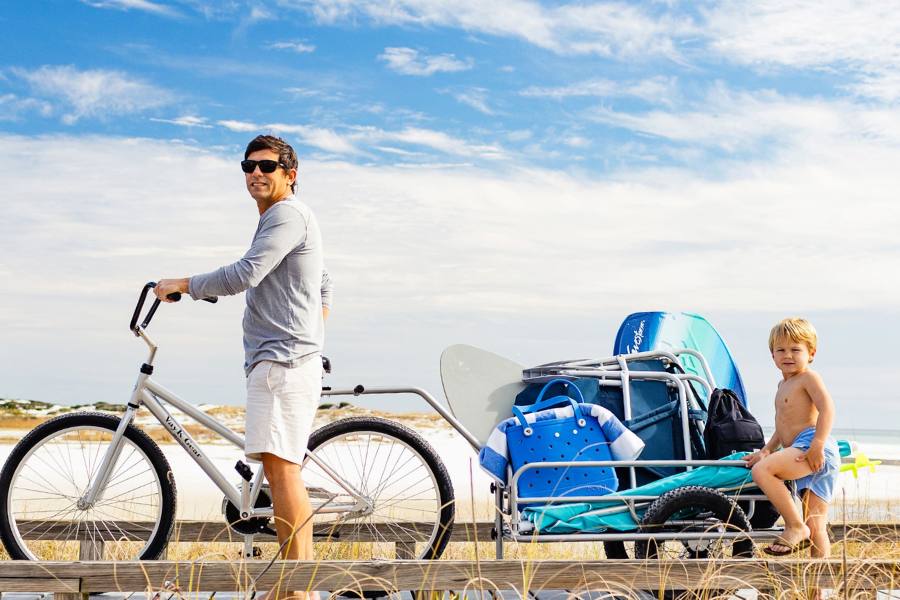 Man riding a bicycle with a kid in a wagon behind him on the beach